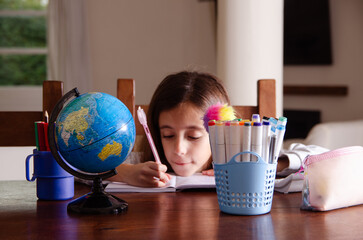 A young girl doing her homework on a wooden table surrounded by school supplies. 