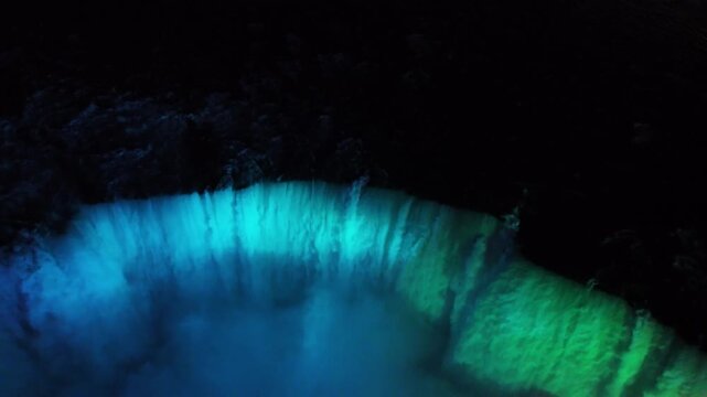 Drone starts at the cliff edge of Horseshoe Falls with illuminated currents, moves into the glowing mist, then pulls out and rises to reveal the Trans-Canada Trail viewpoint and cruises nearing the fa
