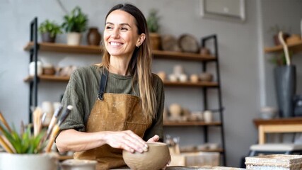 smiling senior woman sculpting a clay bowl in a cozy pottery studio, wearing a brown apron over casual clothes, focused and joyful expression, surrounded by ceramic tools and shelves filled with handm