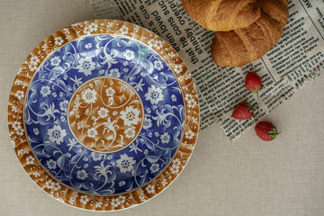 Classic breakfast flat lay. An empty vintage plate with blue and brown floral patterns, with croissants and strawberries on newspaper.