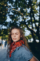 young woman with eyes closed wearing red scarf and denim jacket outdoors in warm sunlight under...