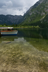 A boat rests on the calm, clear waters of a lake, surrounded by towering, lush green mountains under a cloudy sky.