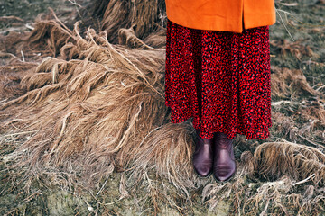 A close-up of boots and red skirt over tangled dry grass and mud