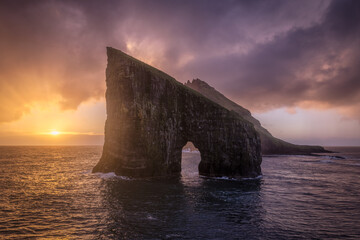 View of a dramatic sea arch carved into a towering cliff face, illuminated by the warm glow of the setting sun, VÃ¡gar, Faroe Islands.