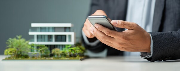 A person in business attire uses a smartphone near a modern architectural house model on a desk.