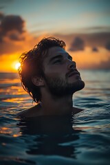 Young Mediterranean man with beard swimming in ocean at sunset, dramatic golden light illuminates profile against dark water and vibrant sky background.