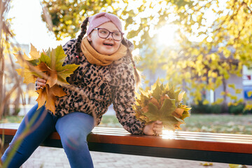 Joyful girl with Down syndrome playing with autumn leaves on bench