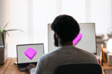 An African American man works on his laptop and external monitor, both displaying a pink 3D cube. He is focused on his creative or technical design project.