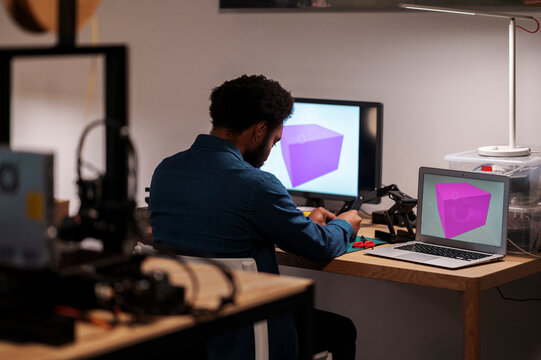 An African American man works on a 3D design project in his workshop. He manipulates a component, with two computer screens displaying a purple cube model.