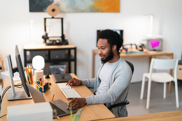 An African American man works diligently at his modern office desk. He uses a laptop, external monitor, keyboard, and trackpad, focused on his tasks in a creative workspace.