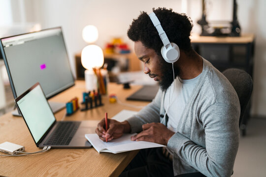 An African American man with headphones on is diligently writing in a notebook at his home office desk. A laptop and large monitor are also present, indicating a productive work or study session.