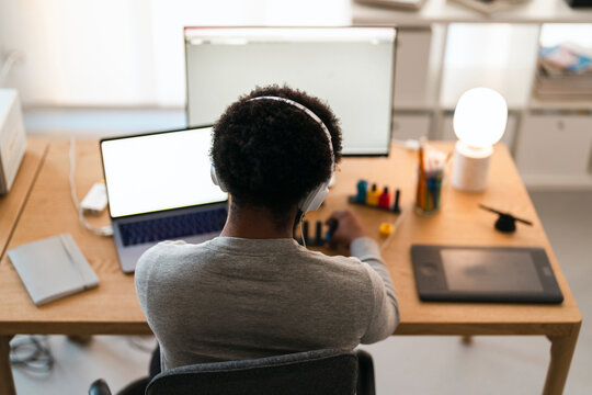 An African American person wearing headphones works at a desk with a laptop and monitor. They are focused on a creative task, using digital tools.