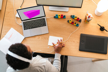 An African American person works at a desk, focused on 3D design on a laptop. They use a pencil and pad, surrounded by creative tools like modular controllers and a graphics tablet.