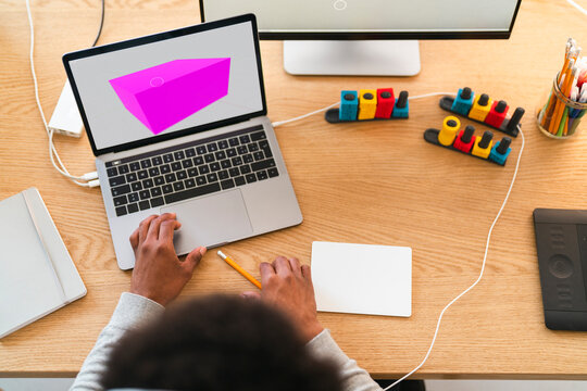 An African American person works on a laptop, creating a 3D model. Their hands are on the trackpad, surrounded by creative tools like a drawing tablet and modular controllers.