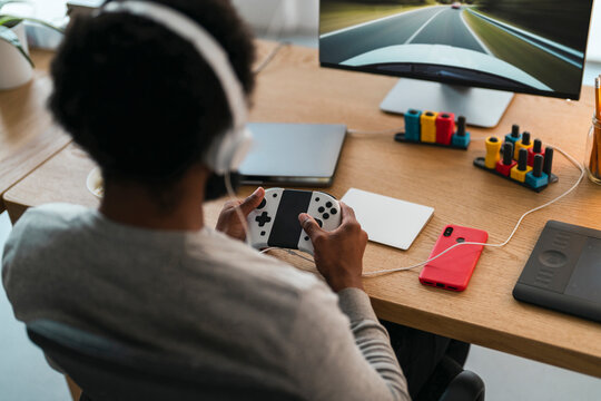 An African American person wearing white headphones is engrossed in a video game. They hold a controller, playing a racing game displayed on a large monitor at their desk.