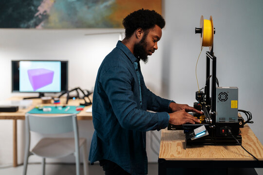 An African American man operates a 3D printer in a modern workshop. He is focused on the machine, with a 3D model visible on a screen in the background.