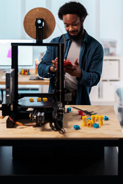 An African American man operates a 3D printer in a modern workshop. He uses his smartphone to control the device, surrounded by colorful 3D printed objects.
