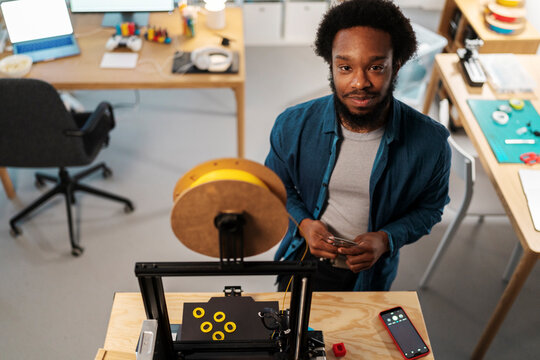An African American man stands in a creative workspace, holding a smartphone. He looks at the camera while a 3D printer with yellow objects operates in front of him.