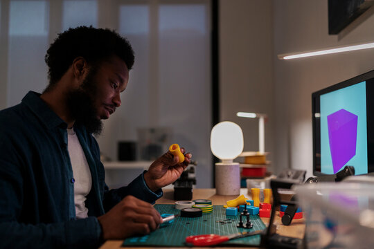 An African American man is focused on assembling a yellow 3D printed object at his desk. He works with various colorful components and tools, with a 3D model on his computer screen.