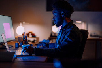 An African American man works intently on his laptop and large monitor in a dimly lit home office. He is focused on his task, illuminated by screen light.