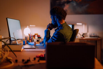 An African American man works at his computer in a dimly lit office. He is focused on the screen, deep in thought, working late into the evening.