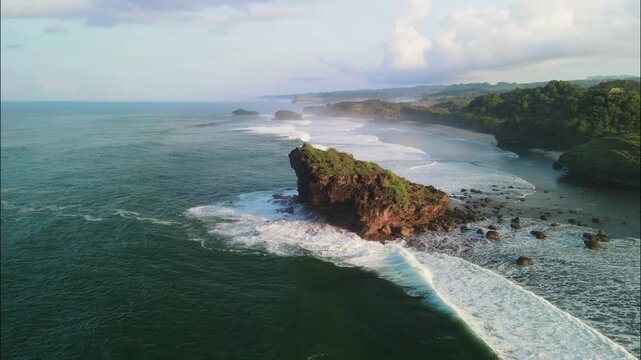 From a cliff-top viewpoint, this stunning aerial shot captures a crescent beach and a bay dotted with numerous green, rocky sea stacks and crashing waves. Aerial Watu Karung beach in Pacitan.