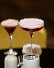 A richly colored red cocktail with a vibrant orange peel garnish is presented in a textured rocks glass, surrounded by other sophisticated drinks in a dimly lit bar setting.