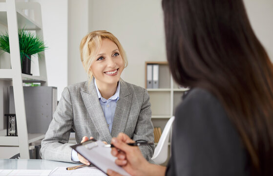 Two businesswomen sitting at office desk and talking about project tasks. Smiling female colleagues collaborating and exchanging ideas while working together on assignments during busy workday. - Powered by Adobe