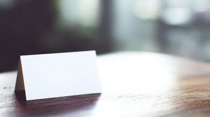 Minimalistic blank white name card propped in A-shape on warm brown wooden table with smooth texture, muted gray and green out-of-focus background, natural indoor lighting.