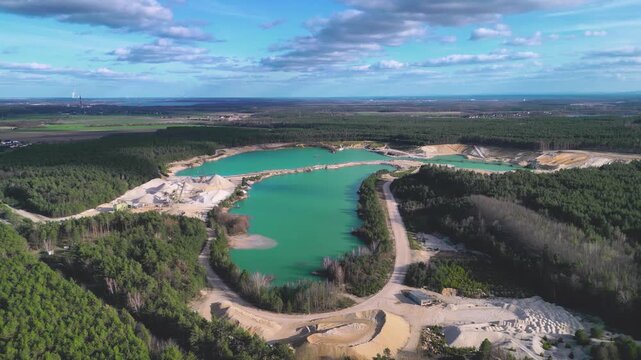 A drone shot of a sand quarry filled with turquoise water, surrounded by forest near the horizon. The open-pit mine with clear blue lakes, sand dunes, and industrial equipment creates a unique 