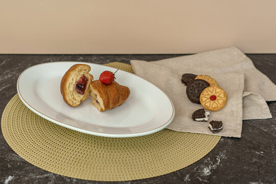 Sweet breakfast still life. A jam-filled croissant topped with a strawberry on a plate, with a side of assorted cookies on a napkin.