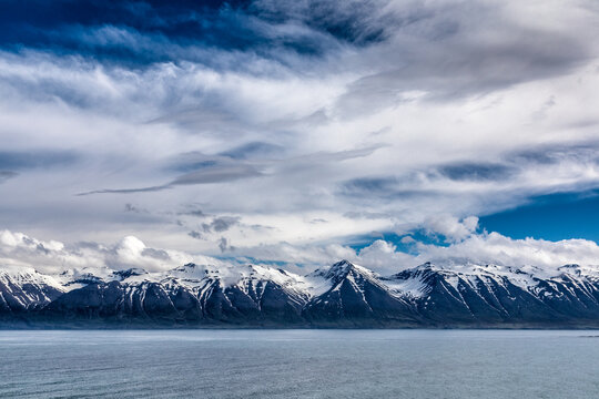 View of snow-capped mountains meeting the tranquil, glassy waters under a dramatic sky filled with swirling clouds, H&Atilde;&para;fn, Iceland.