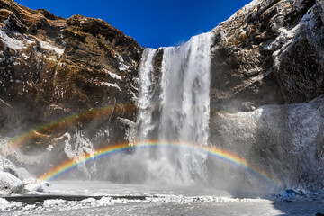 View of a majestic waterfall plunges into a misty pool, crowned by a vibrant rainbow arching across the scene against a backdrop of rugged cliffs, Iceland.