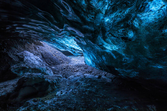 View of an ethereal ice cave, illuminated with vibrant blue light cascading over the rugged, textured ice formations, creating a mesmerizing spectacle, Iceland.