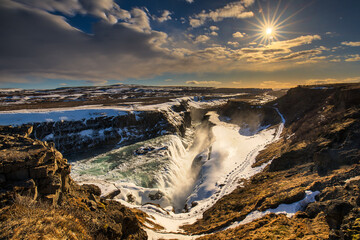 View of Gullfoss waterfall plunges into a rugged canyon, a spectacle of nature's power under a radiant sun and dramatic Icelandic sky, Iceland.