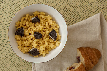 Top view of a breakfast bowl with star-shaped honey cereal and chocolate sandwich cookies. A croissant rests on a napkin. Fun kids breakfast idea.