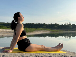 Adult woman sitting on yellow exercise mat and doing yoga outdoors by lake and forest. Summer...