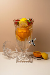 Elegant crystal glass beverage dispenser with tap, filled with refreshing infused detox water with orange, lemon, strawberry, and mint leaves.