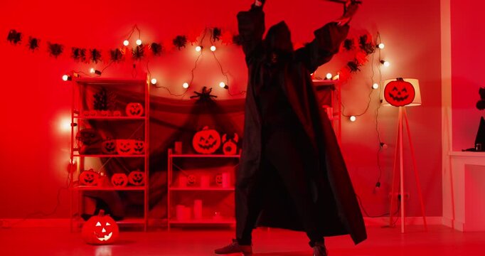 Mysterious figure in dark hooded cloak dancing with scythe in eerie red-lit room decorated with glowing jack-o-lanterns and garlands. Funny person in grim reaper death costume celebrating Halloween.
