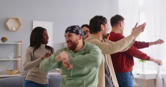Group of carefree positive friends have fun at home and dance to music, enjoy spending time together, celebrate birthday, move actively to favourite song. Friendship and emotions concept