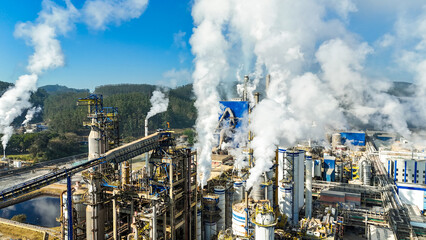 Indústria de papel vista do alto com várias chaminés e caldeiras em funcionamento. A imagem destaca a emissão de fumaça e o contraste entre produção e natureza ao redor