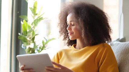 Cheerful woman browsing digital tablet while lounging comfortably, connecting online through social network, enjoying leisure or professional activities - Powered by Adobe