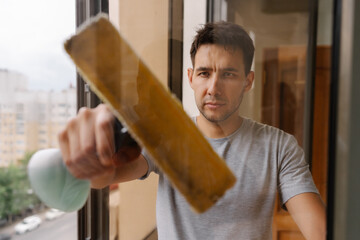 Focused young man washing window using squeegee and detergent, ensuring spotless and streak-free...