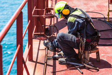 A crew member working on the deck of a merchant ship, using a pneumatic tool to remove rust.