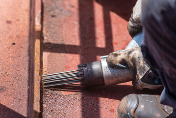 Worker wearing gloves uses a pneumatic tool to remove rust from a metal surface, likely on a ship....