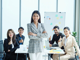 Confident Asian businesswoman standing with arms crossed while diverse business team shows support with thumbs up, representing leadership and team unity in modern office.