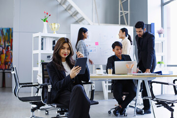 Confident Asian businesswoman using tablet while coworkers collaborate in modern office setting with creative brainstorming and digital tools around.