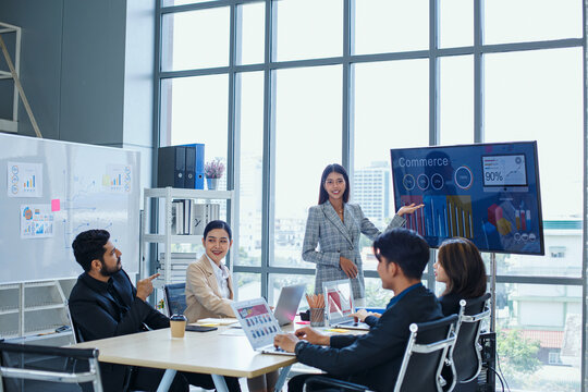 Asian businesswoman leading corporate presentation to diverse team in high-rise office showing e-commerce data and sales growth on digital display screen.