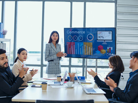 Professional Asian and multicultural team discussing performance metrics during formal meeting in office with digital marketing analytics on large screen.