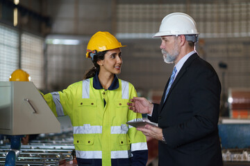 Fototapeta premium A foreman and a woman worker are standing in a factory, looking at a tablet. The engineer talks with the woman's workers. They wear a yellow and white safety helmet for safety.